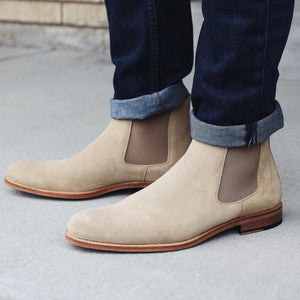Close-up of a person wearing beige suede Chelsea boots and dark blue jeans with cuffs, standing on a concrete surface.
