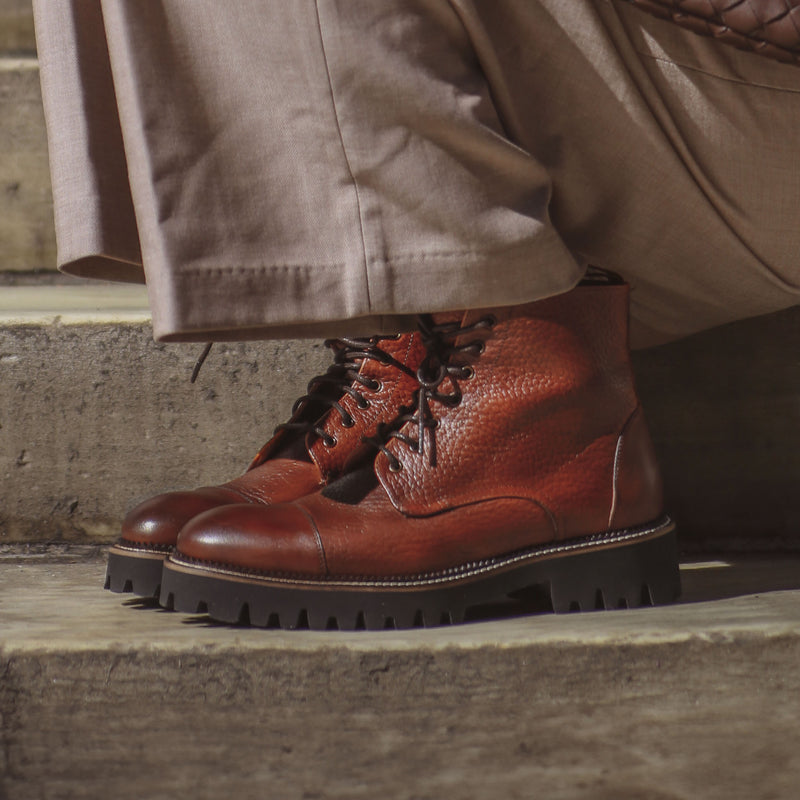 Close-up of a person wearing brown leather lace-up boots with thick tread soles, paired with beige trousers, standing on stone steps.
