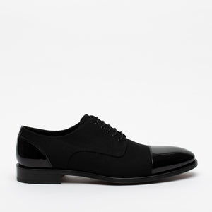A single black oxford shoe with a polished cap toe and heel. The shoe has black laces and a low heel. The background is plain white.