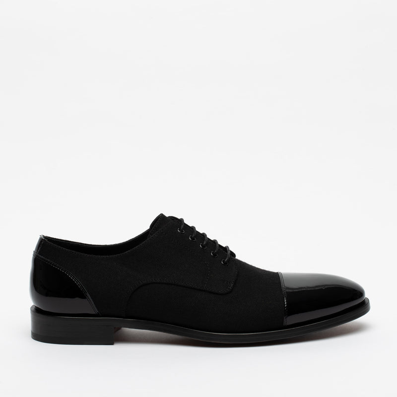A single black oxford shoe with a polished cap toe and heel. The shoe has black laces and a low heel. The background is plain white.