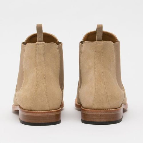 Rear view of a pair of beige suede Chelsea boots with tan soles and pull tabs.